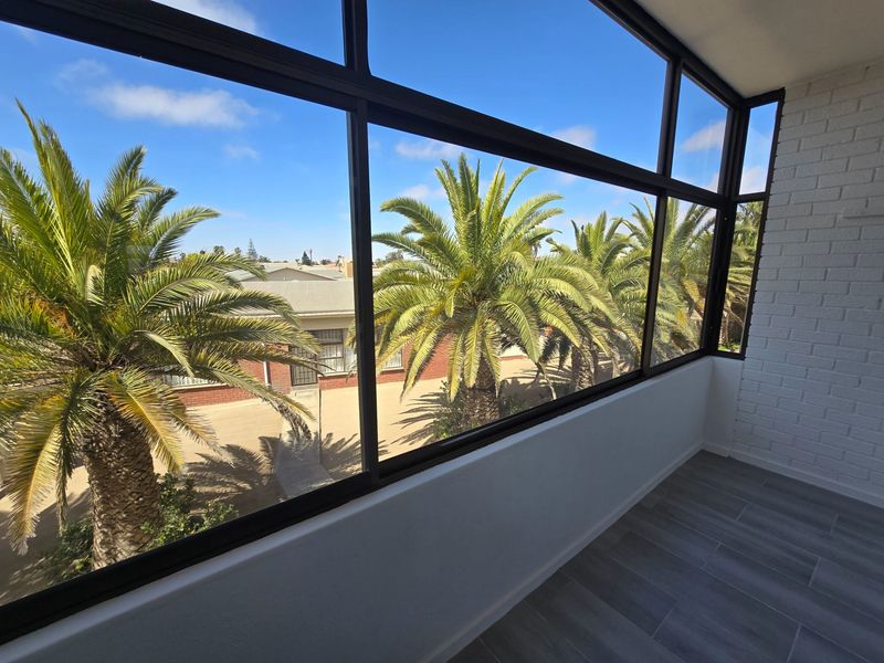 A bright, enclosed balcony featuring large windows with views of palm trees, finished with modern tiled flooring and a painted brick wall.