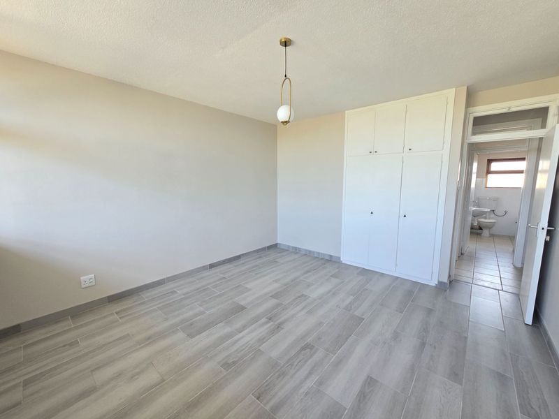 A bright, empty bedroom featuring modern wood-look tiled flooring, built-in storage cupboards, and a contemporary pendant light fixture.