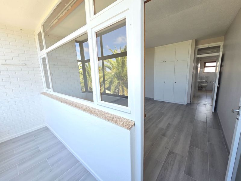 An enclosed balcony area featuring modern grey tiled flooring, a white brick wall, and built-in storage cupboards, leading into the main apartment interior.