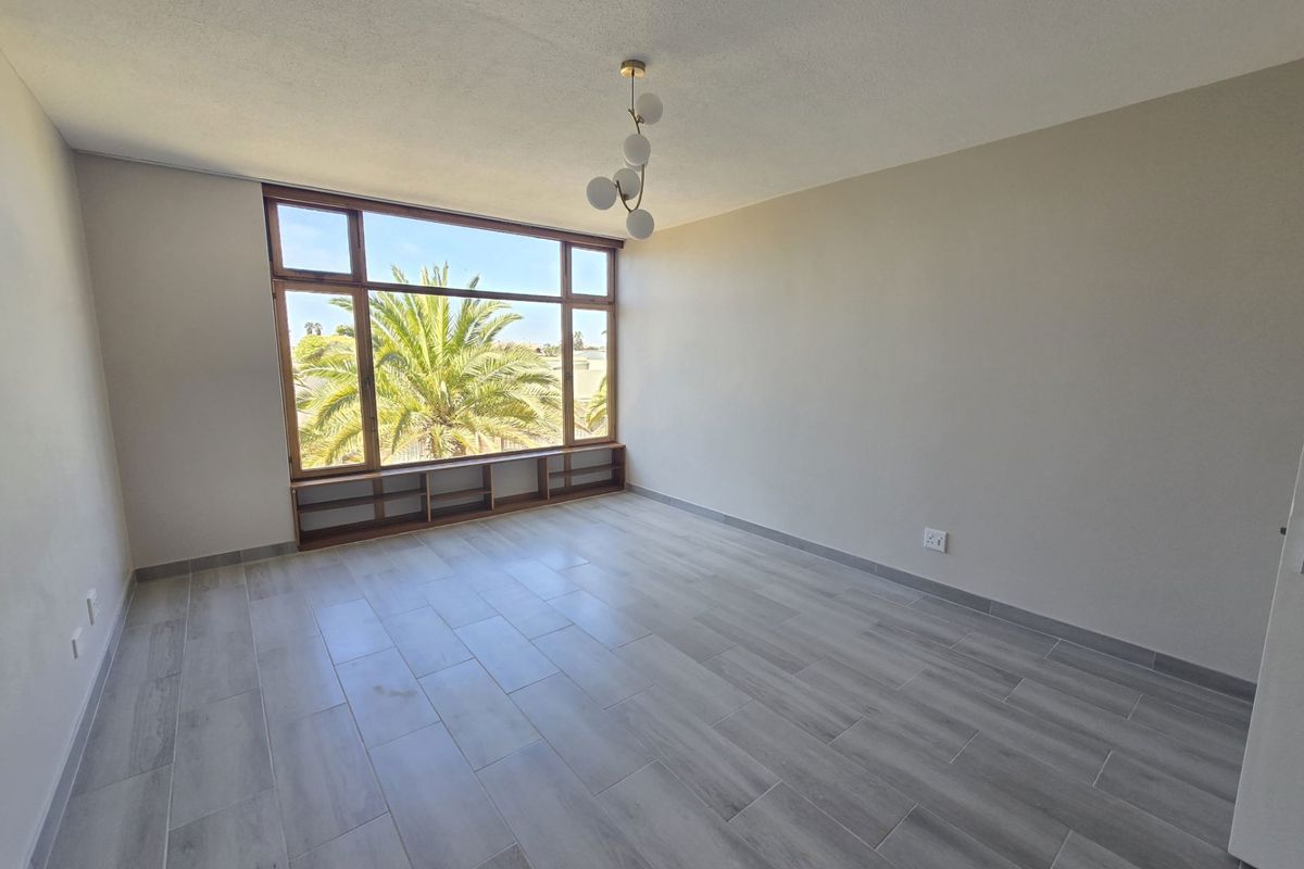 A bright, empty bedroom featuring modern grey tiled flooring, a large window with built-in low shelving, and a contemporary light fixture.