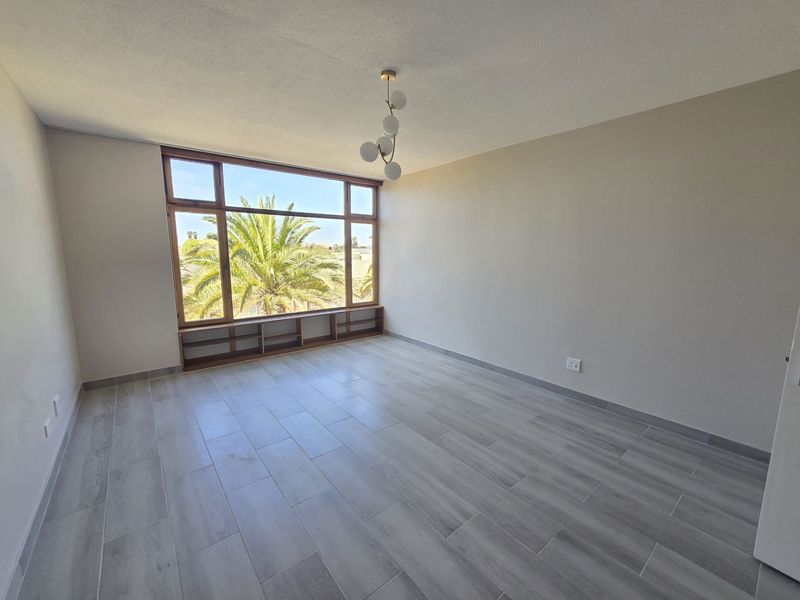 A bright, empty bedroom featuring modern grey tiled flooring, a large window with built-in low shelving, and a contemporary light fixture.