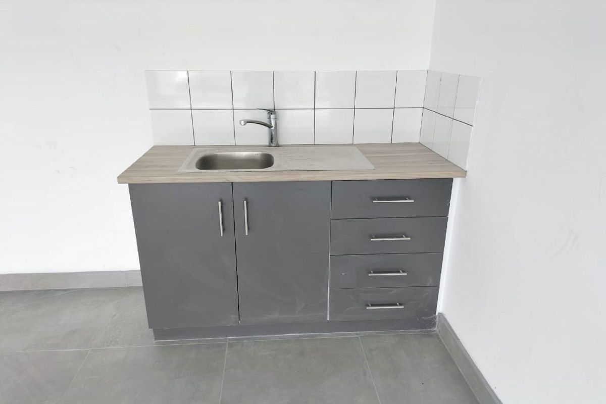 A compact kitchenette featuring grey cabinetry, a stainless steel sink with mixer tap, laminate countertop, and a clean white tiled splashback.