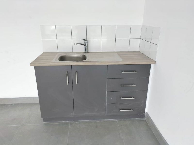 A compact kitchenette featuring grey cabinetry, a stainless steel sink with mixer tap, laminate countertop, and a clean white tiled splashback.