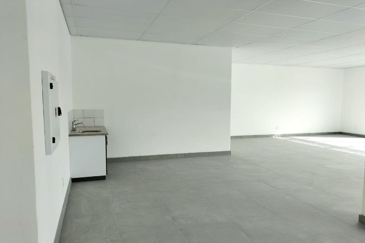 A spacious, open-plan retail or office area featuring neutral tiled flooring, a drop ceiling, and a compact kitchenette corner with a sink.