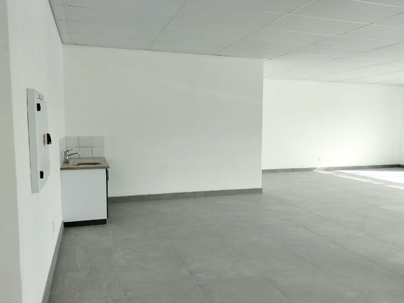 A spacious, open-plan retail or office area featuring neutral tiled flooring, a drop ceiling, and a compact kitchenette corner with a sink.