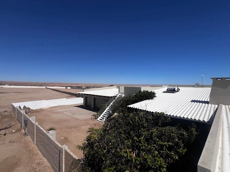 Aerial view of the property showing the main building with a corrugated metal roof, solar water heater, and surrounding perimeter wall in a desert landscape.
