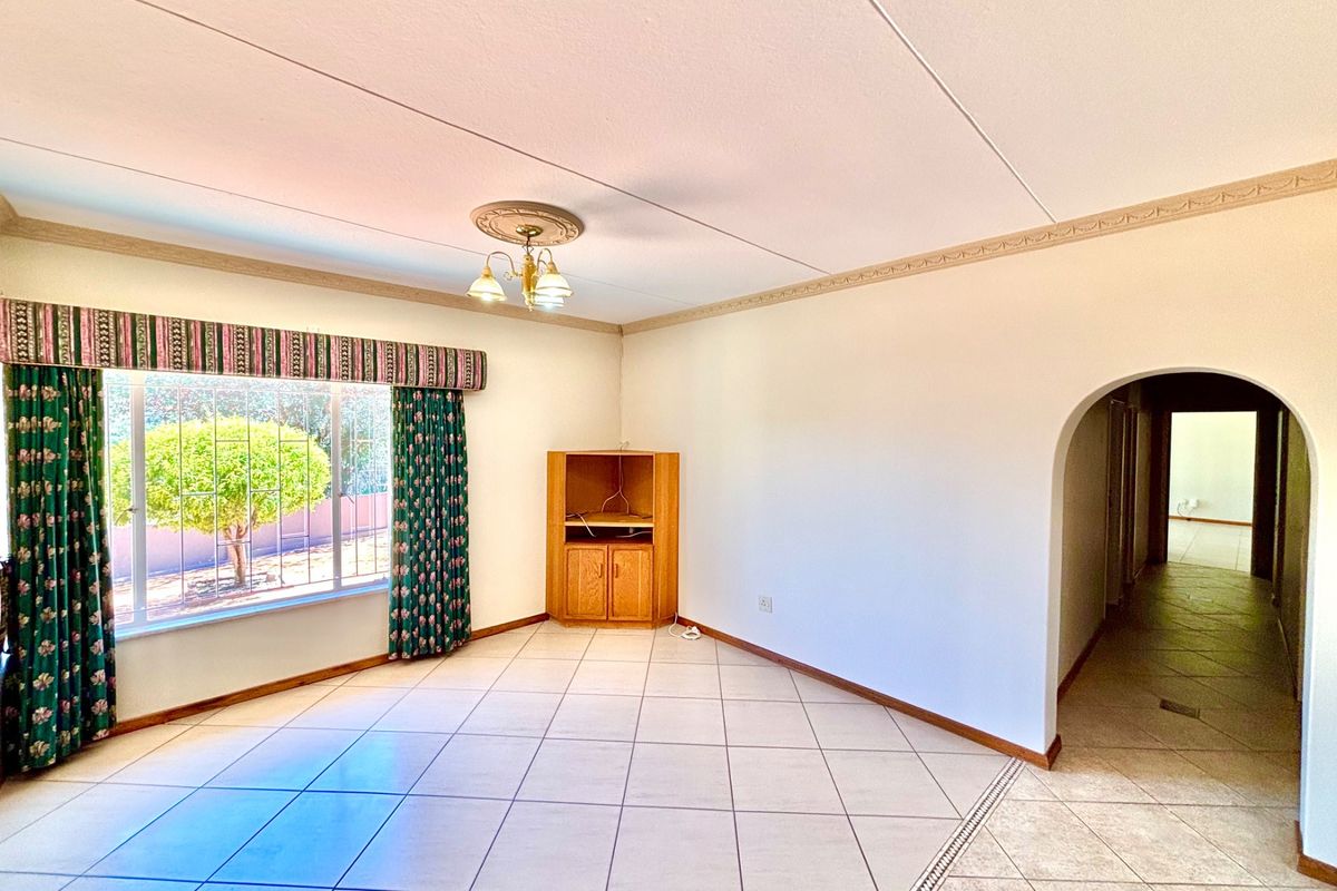 A spacious, light-filled living room featuring neutral tiled flooring, a decorative ceiling rose, and a classic arched entryway leading to the hallway.