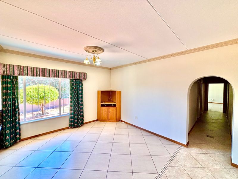 A spacious, light-filled living room featuring neutral tiled flooring, a decorative ceiling rose, and a classic arched entryway leading to the hallway.