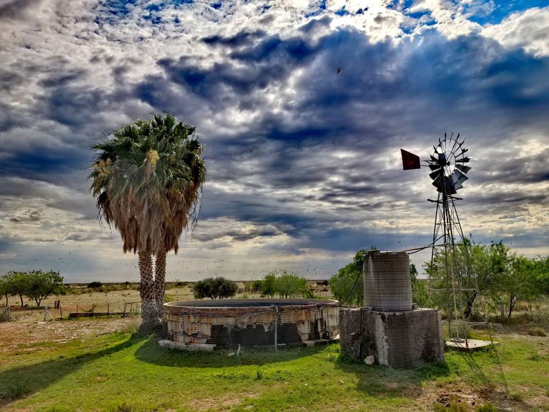 A rural farm landscape featuring a traditional windmill, a water reservoir, and a storage tank set against a dramatic cloudy sky.