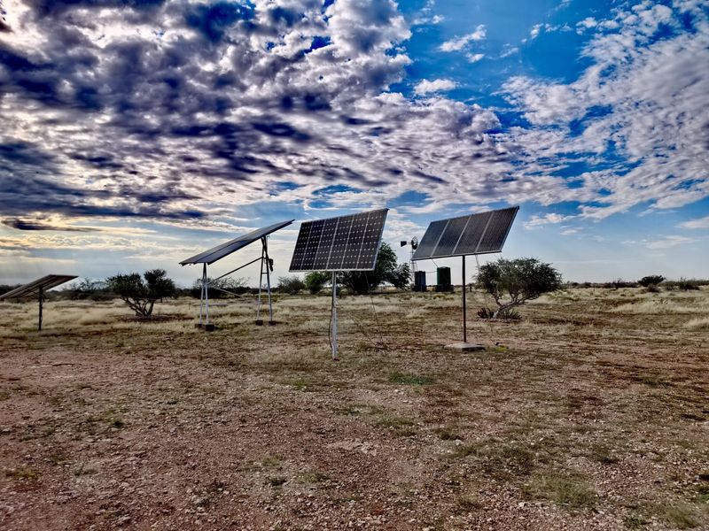 Open farm land featuring ground-mounted solar panel arrays, a traditional windmill, and water storage tanks under a dramatic sky.