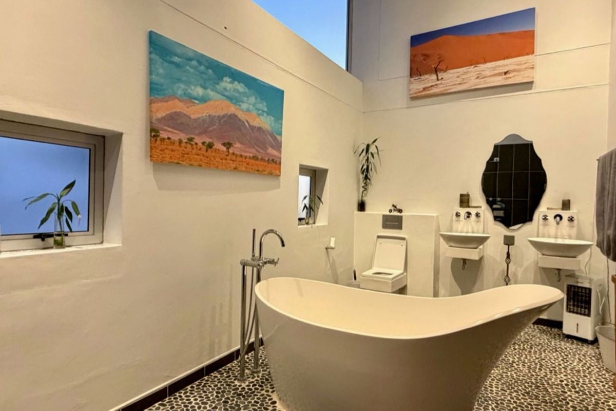 Modern bathroom featuring a freestanding bathtub, dual wall-mounted sinks, wall-mounted toilet, and unique pebble tile flooring with high ceilings and natural light.
