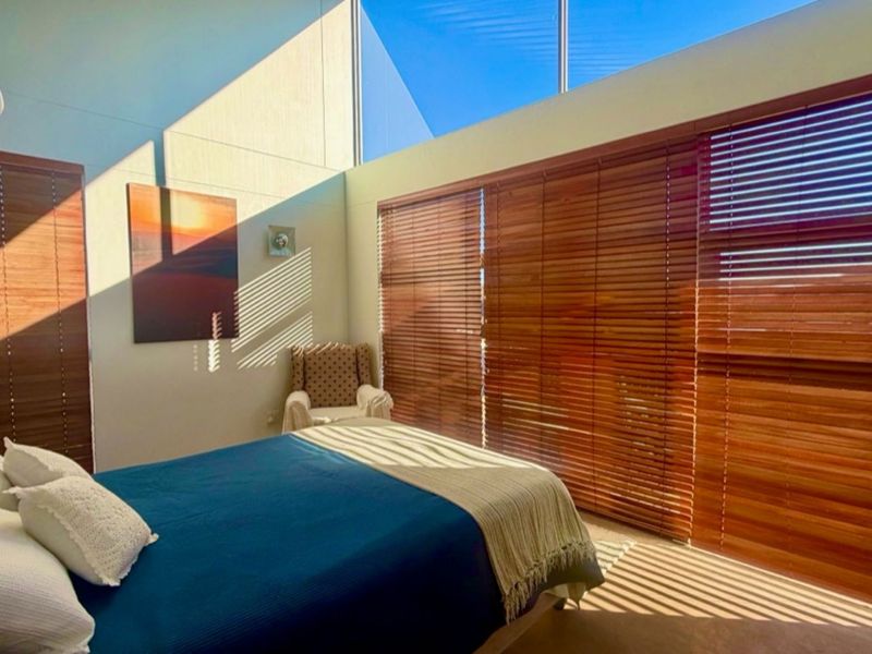 A bright bedroom featuring a high sloped ceiling, clerestory windows for natural light, and large sliding doors with timber venetian blinds.