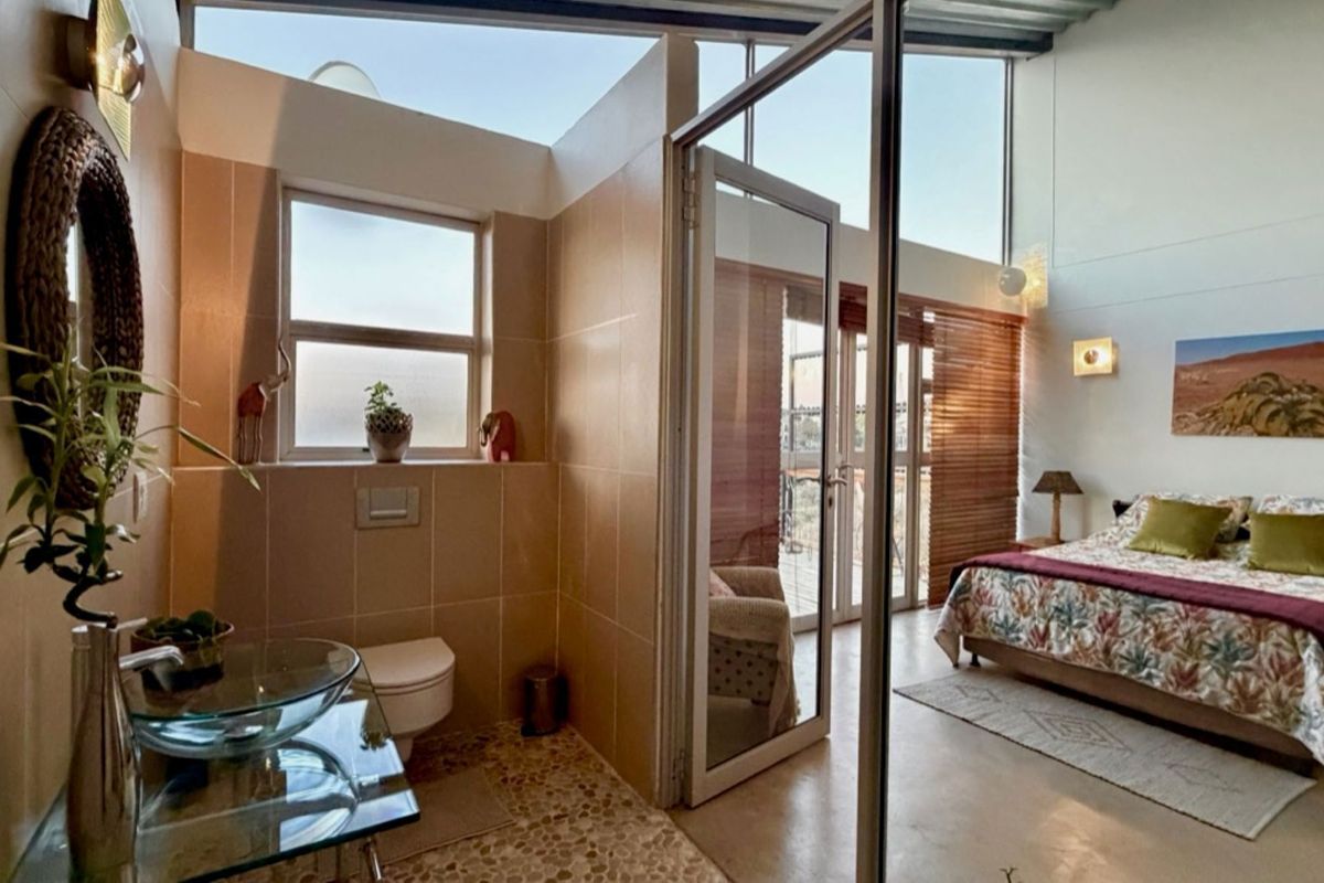 A modern bedroom featuring an open-plan en-suite bathroom with a glass vessel sink, wall-mounted toilet, and large windows providing ample natural light.
