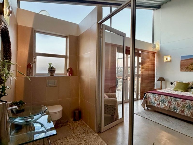A modern bedroom featuring an open-plan en-suite bathroom with a glass vessel sink, wall-mounted toilet, and large windows providing ample natural light.