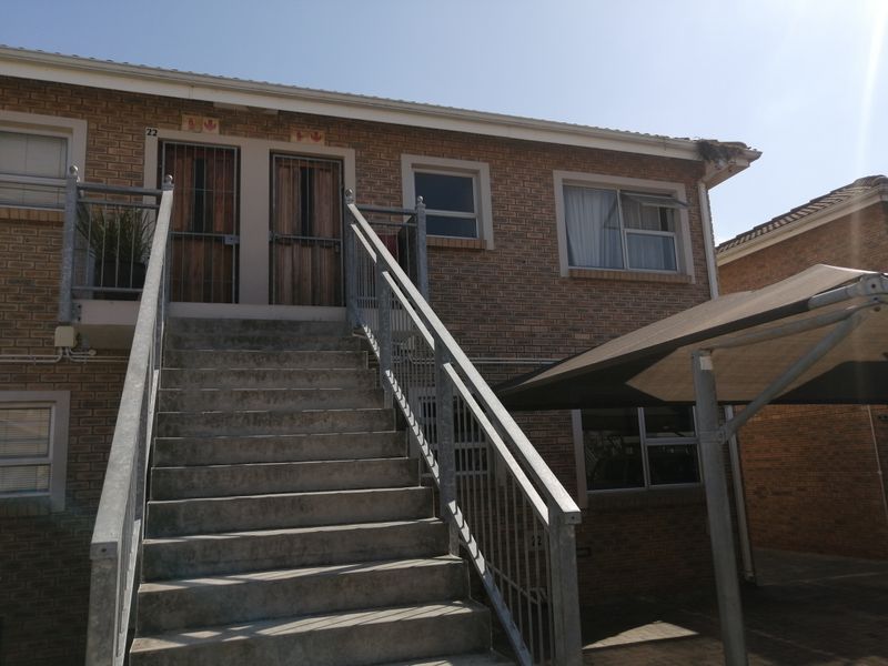 Exterior view of a face brick apartment building featuring an external concrete staircase with metal railings and an adjacent shade cloth carport.