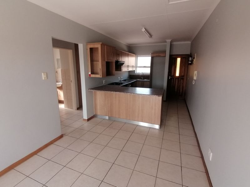 An open-plan kitchen area featuring built-in cabinetry, laminate countertops, and tiled flooring, leading to an adjacent bathroom and main entrance.