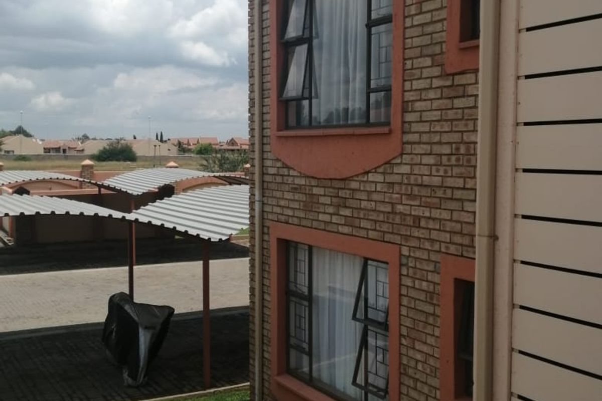 Side view of a double-storey townhouse featuring face brick construction, aluminium window frames, and an adjacent paved area with carport access.