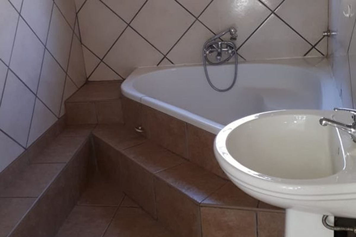 A functional bathroom featuring a corner bathtub with tiled surround, a pedestal basin, and a window providing natural light.