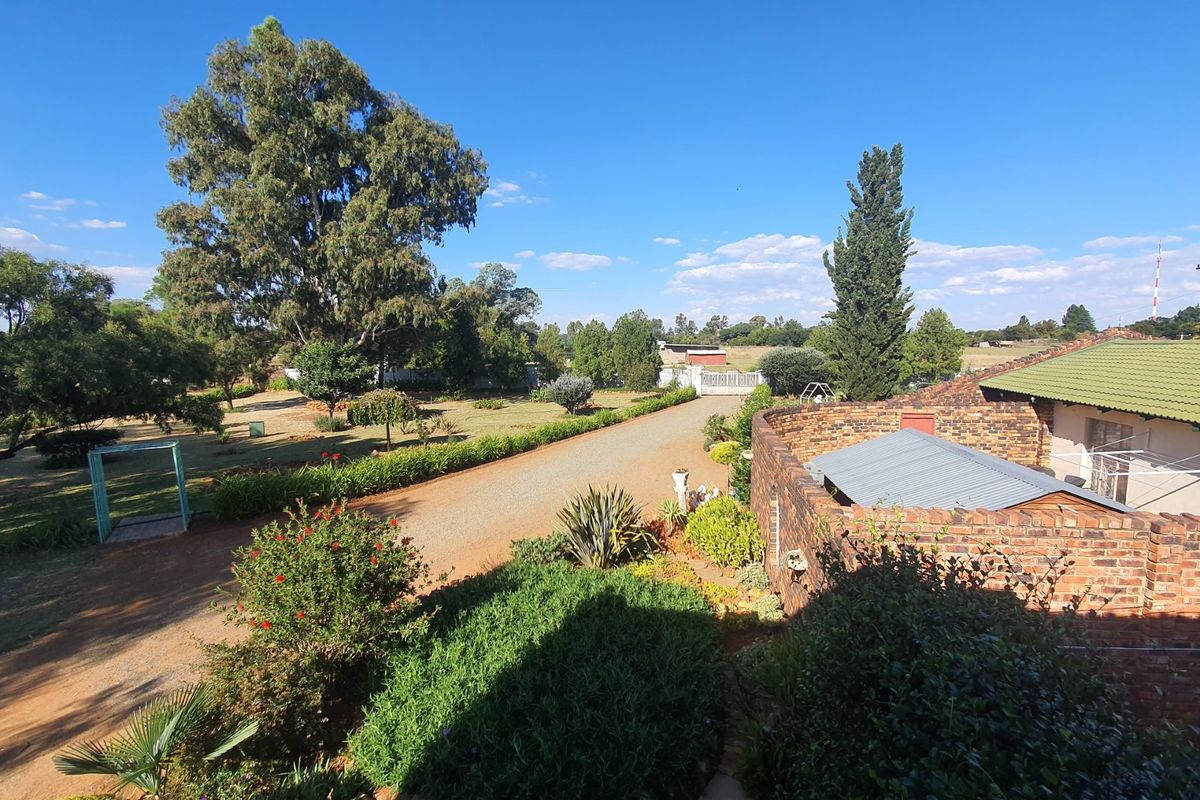 An elevated view of a farm property featuring a brick building with a corrugated roof, a gravel driveway, and expansive landscaped garden grounds.