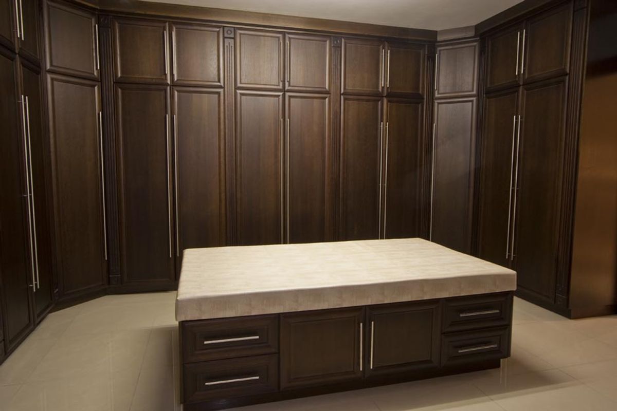 A spacious walk-in dressing room featuring extensive dark wood cabinetry, floor-to-ceiling storage, and a central island with a stone countertop.