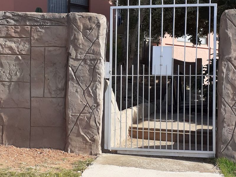 Front entrance view featuring a metal security gate set within a textured concrete boundary wall and a concrete pathway leading to the property.