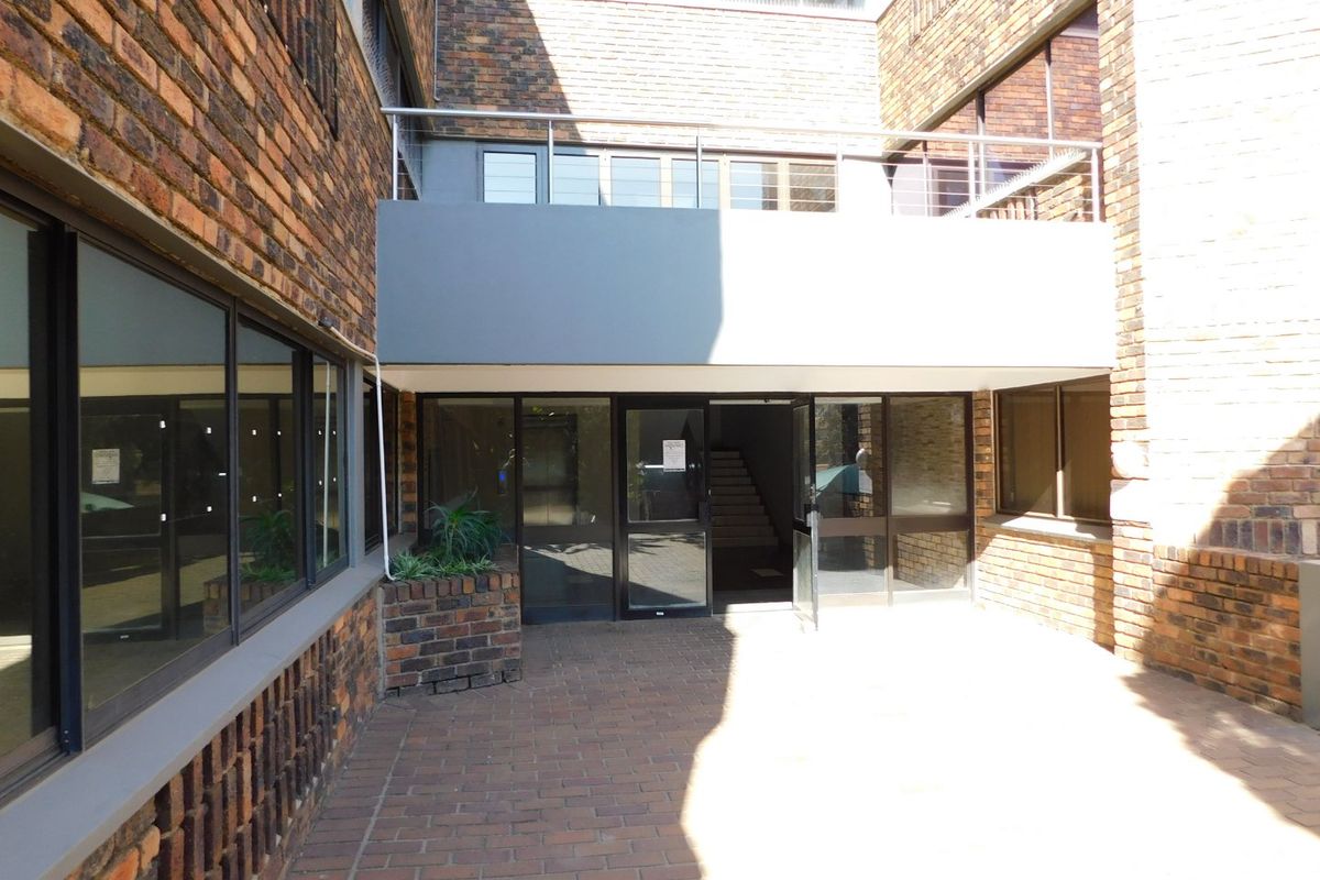A paved courtyard area featuring a brick facade, aluminium framed windows, and a balcony with metal railings, providing access to the office building entrance.