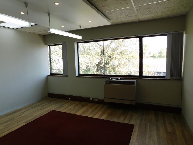 An empty office space featuring laminate flooring, a suspended ceiling with integrated lighting, and a wall-mounted HVAC unit with large windows.