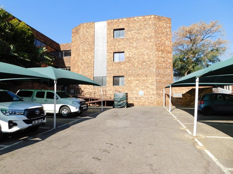 Front exterior view of the office building featuring a brick facade, paved parking area with shade cloth carports, and an accessible wheelchair ramp.