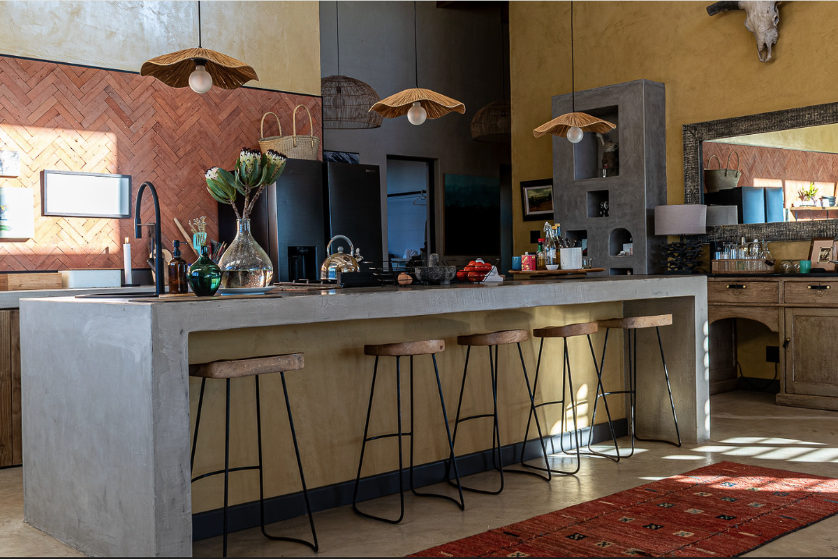 A stylish kitchen featuring a large concrete island, herringbone brick backsplash, and custom built-in concrete shelving with a warm, textured aesthetic.