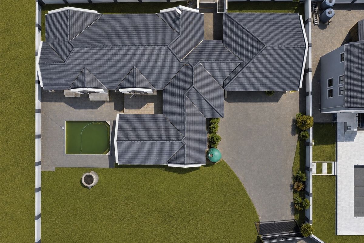 Aerial view of a residential property featuring a large tiled roof, paved driveway, perimeter wall, and a swimming pool requiring maintenance.