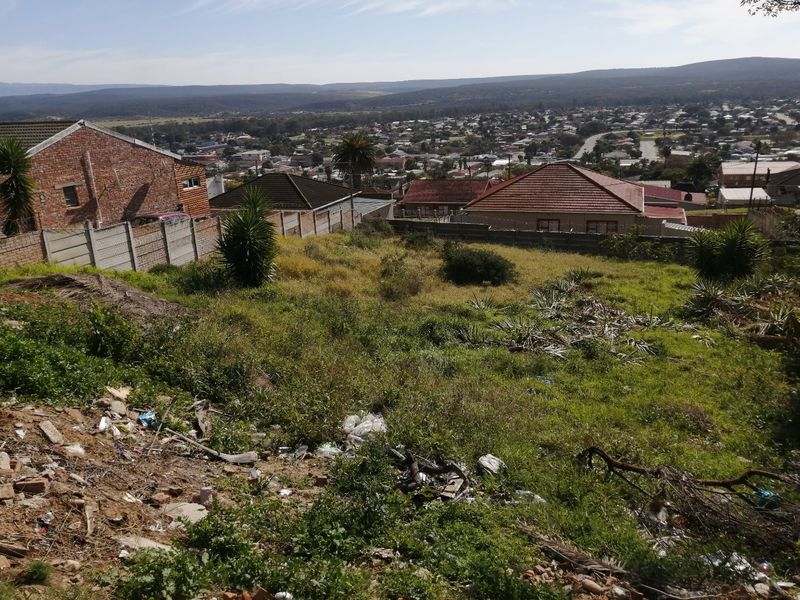 A sloped vacant residential plot featuring overgrown grass and scattered debris, offering elevated views over the surrounding suburban neighborhood.