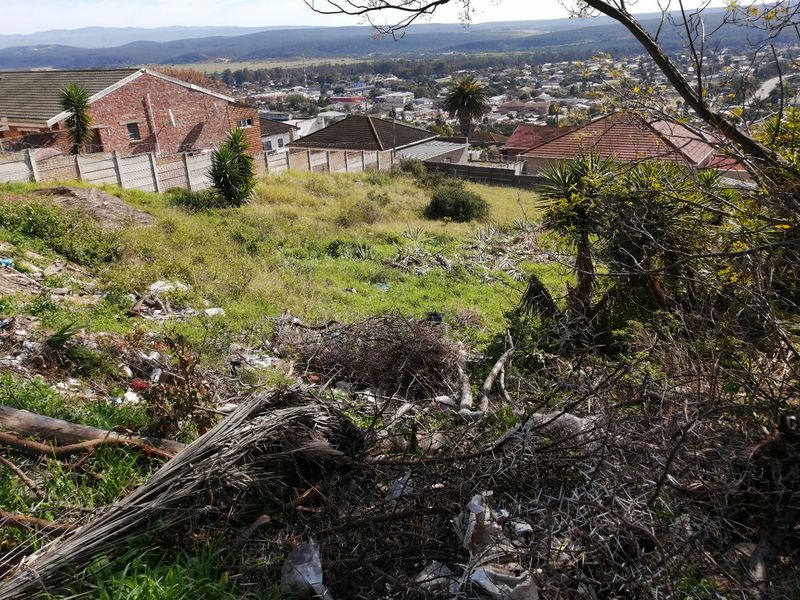 A sloped vacant residential plot featuring natural vegetation and panoramic views of the surrounding neighborhood and distant mountains.