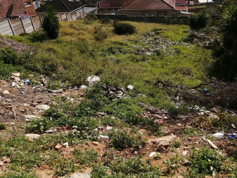 A vacant residential plot featuring a sloped landscape with overgrown vegetation and accumulated debris, enclosed by perimeter boundary walls.