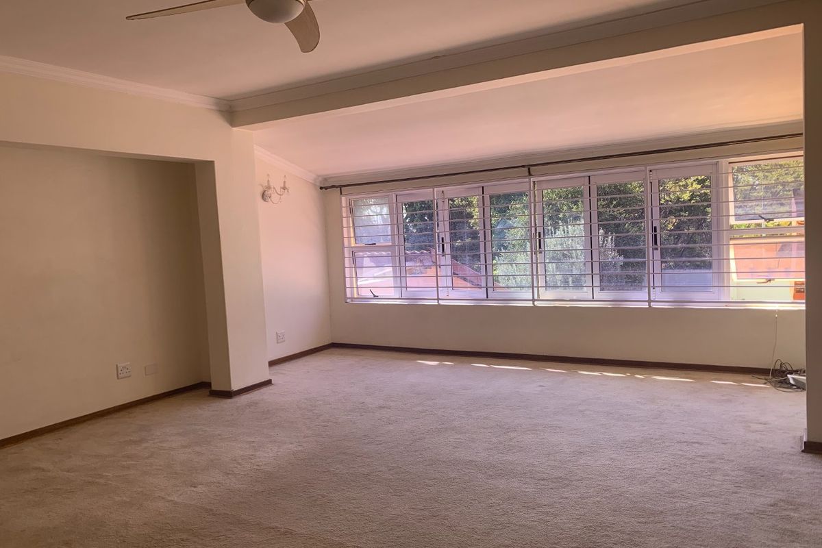Spacious, sunlit living area featuring neutral carpeting, a ceiling fan, and large windows with security bars, offering a bright and open living space.
