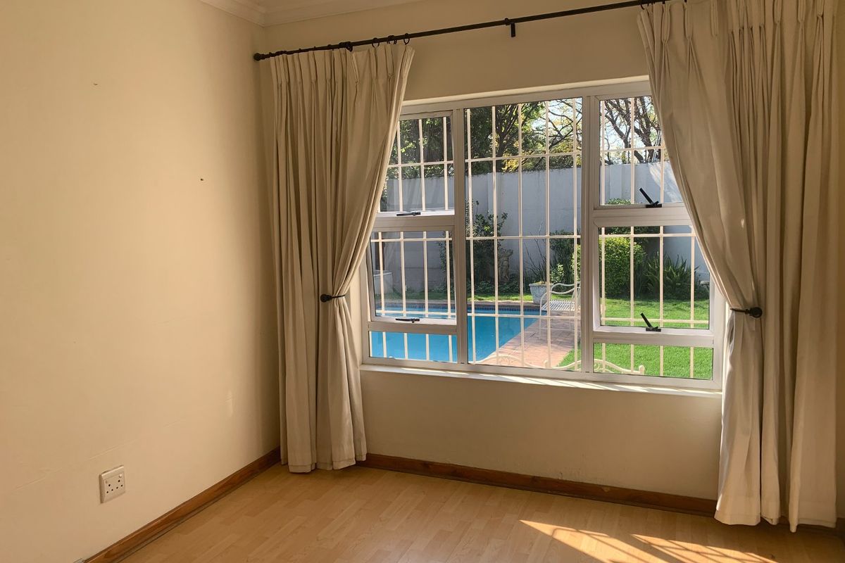 A bright, empty bedroom featuring light wood-look flooring, large windows with security bars, and a view overlooking the garden and swimming pool area.