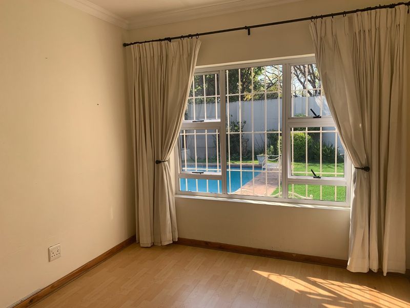 A bright, empty bedroom featuring light wood-look flooring, large windows with security bars, and a view overlooking the garden and swimming pool area.