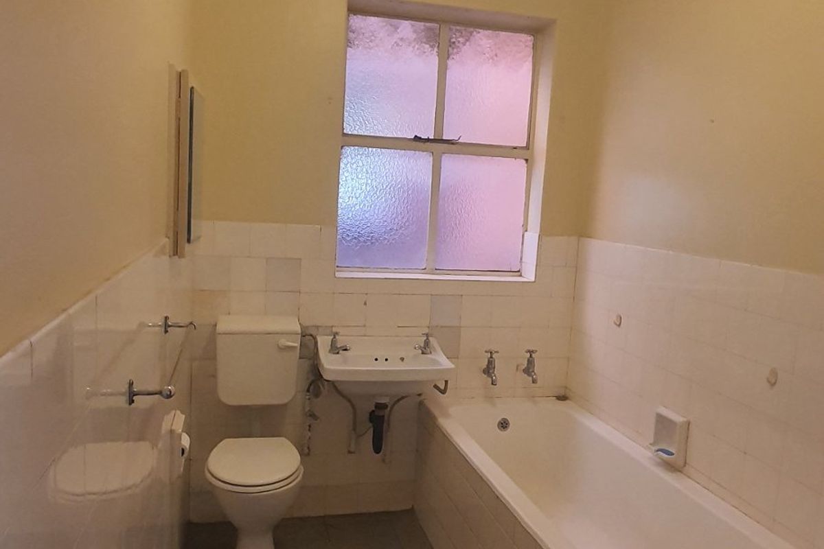 A dated bathroom featuring a standard bathtub, wall-mounted basin, and toilet, with original tiling and a frosted glass window.