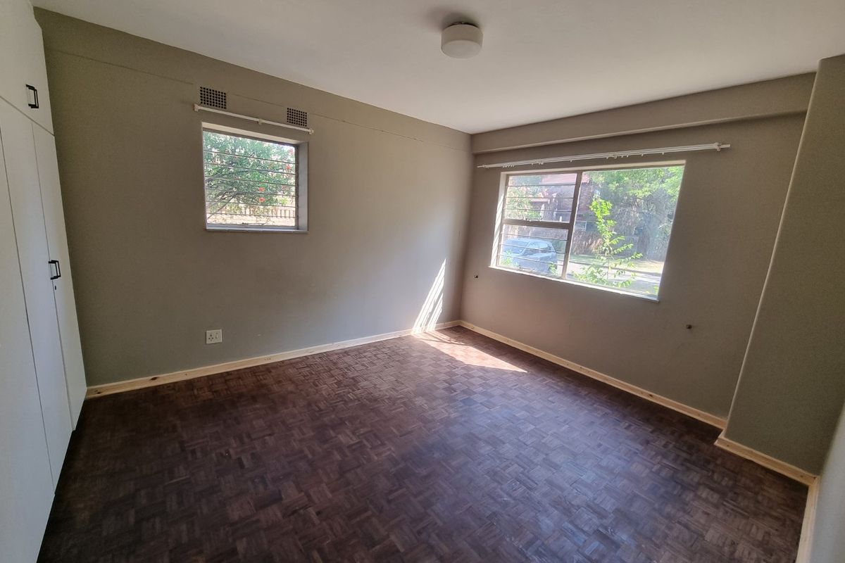 A bright, empty bedroom featuring parquet flooring, built-in cupboards, and large windows with security bars.