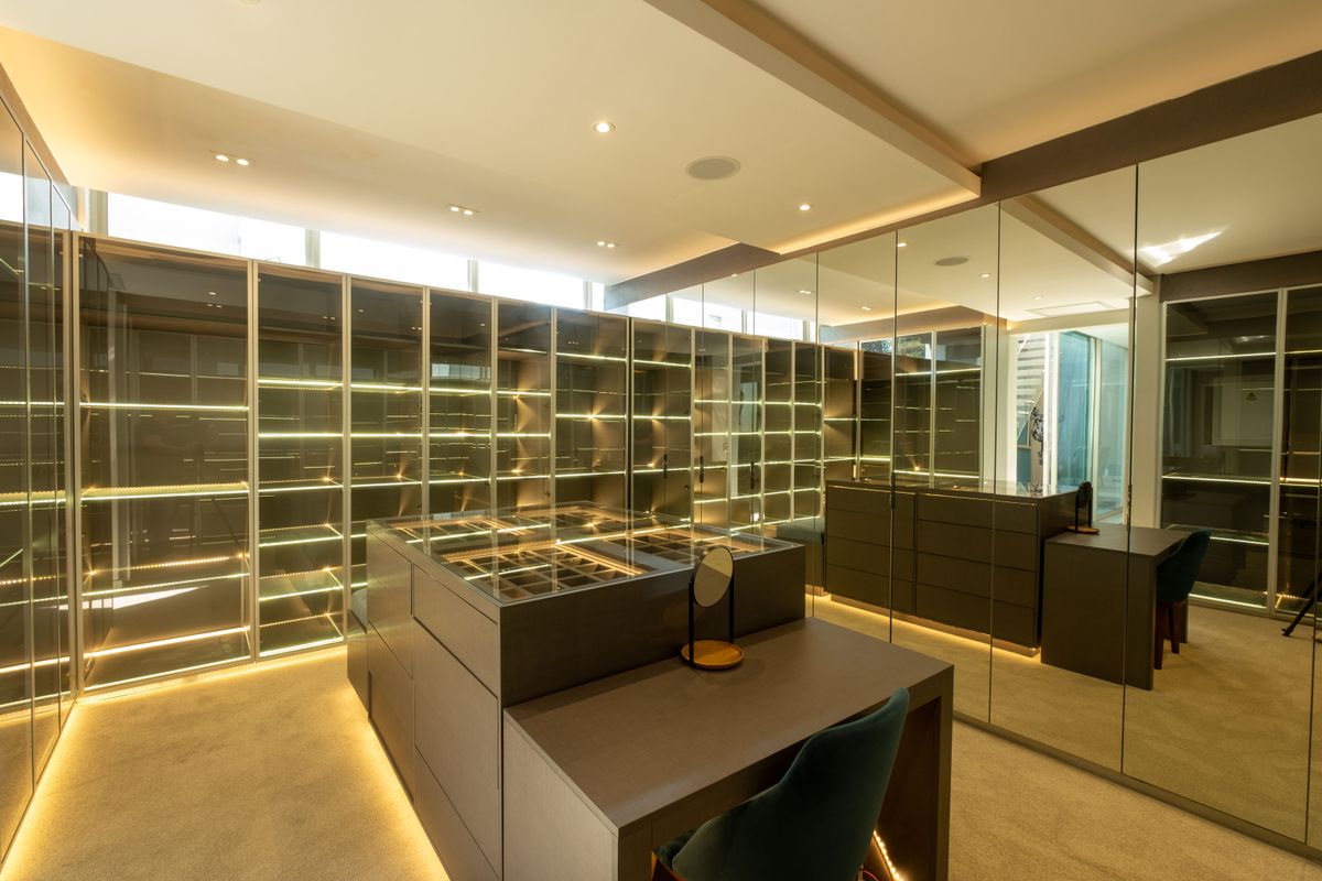 A modern, high-end walk-in dressing room featuring extensive built-in cabinetry, integrated LED shelf lighting, mirrored wardrobe doors, and a central island unit.