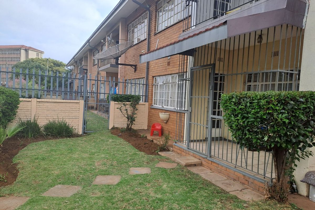 Exterior view of a brick apartment building featuring a ground-floor patio area with security gates and a small private garden space.