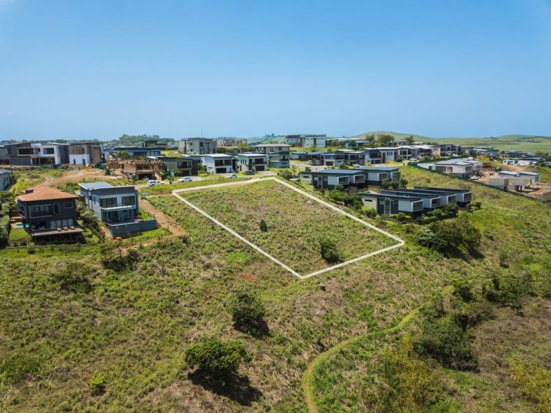 An aerial view of a vacant residential plot situated within a developed estate, featuring sloped terrain and surrounding modern homes.