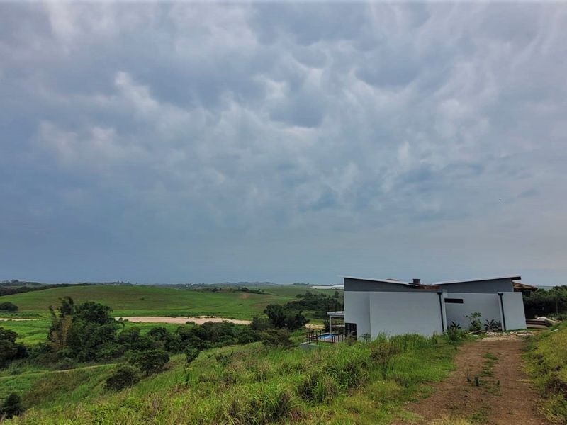 An elevated view of a modern residential property set within a vast, open landscape with rolling green hills and a cloudy sky.