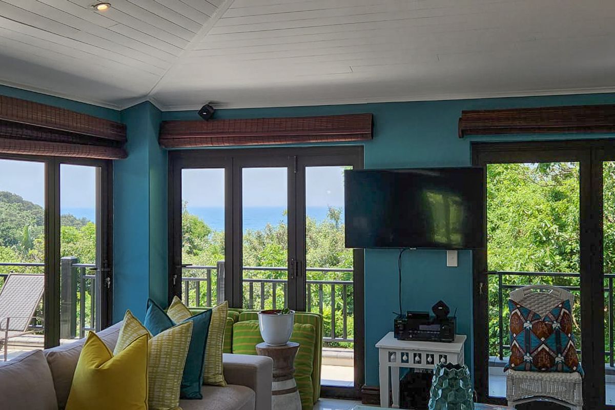 A bright living room featuring high vaulted ceilings, vibrant blue walls, and multiple glass doors opening onto a scenic balcony with ocean views.