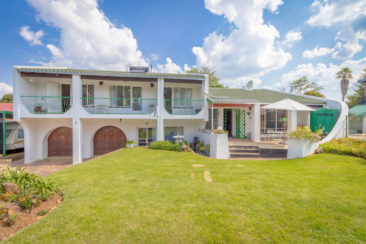 Rear view of a multi-level house featuring a green tiled roof, upper-level balcony, arched garage doors, and a paved patio area overlooking the garden.