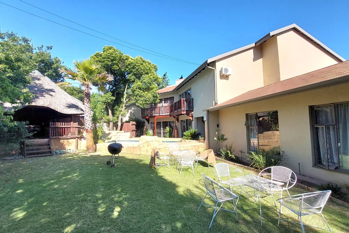 Spacious rear garden featuring a lawn, swimming pool with rock features, a thatched lapa, and a multi-level house with a balcony.