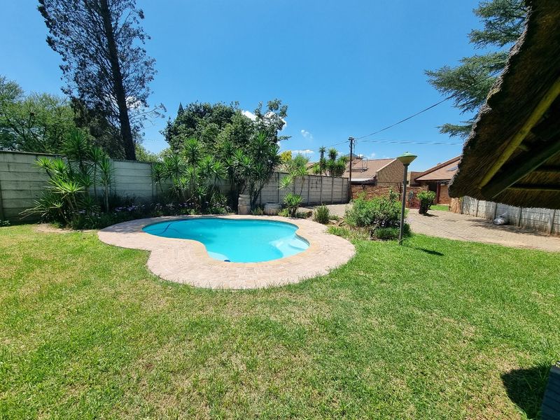 A private backyard featuring a kidney-shaped swimming pool with brick coping, surrounded by a lawn, boundary walls, and a paved driveway area.