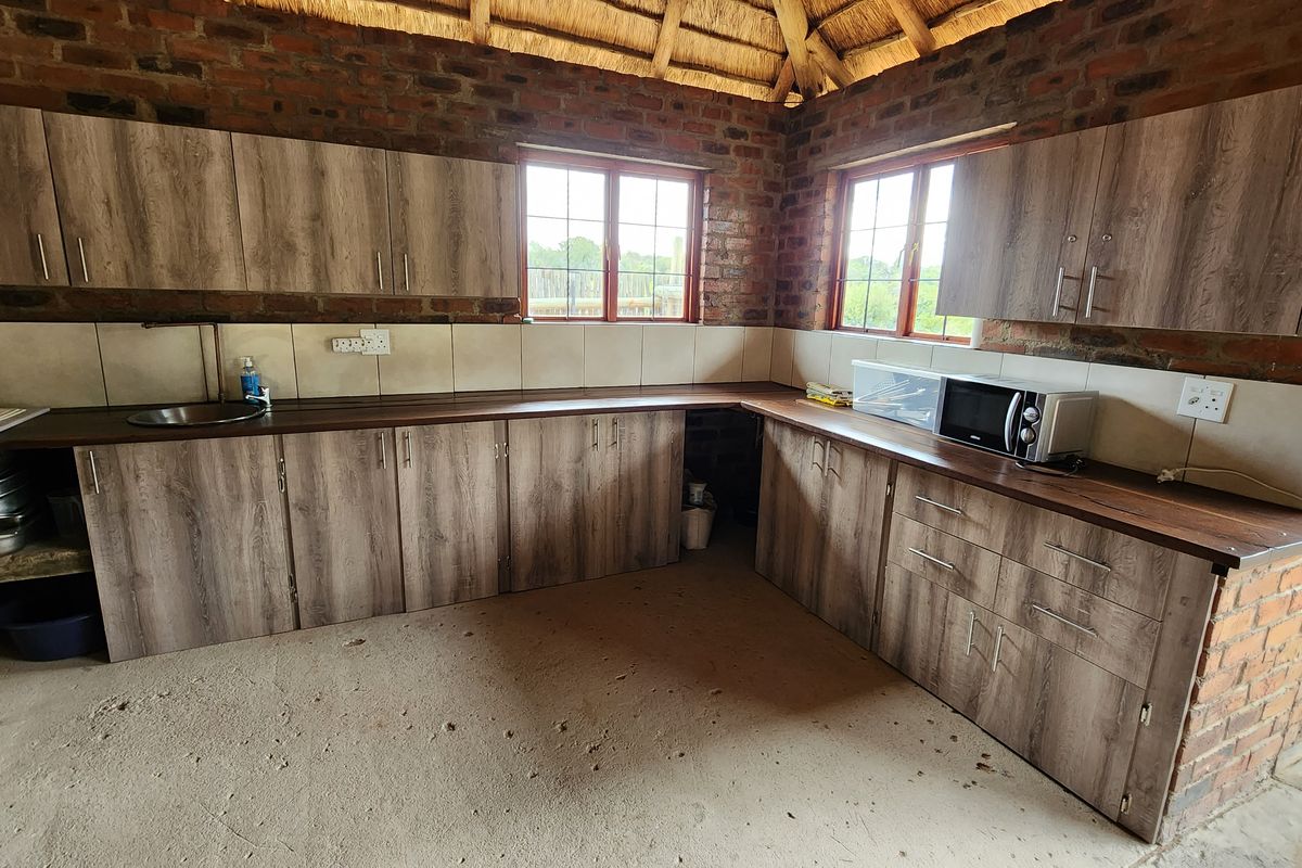 Rustic kitchen area featuring wood-look cabinetry, dark countertops, tiled backsplash, and exposed brick walls under a traditional thatched roof structure.