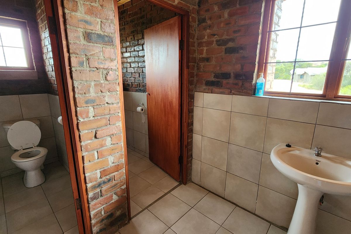 A rustic bathroom featuring exposed brick walls, ceramic floor tiles, a pedestal basin, and a separate toilet cubicle with natural light.