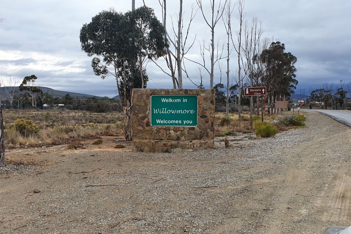 A street-level view of the Willowmore town entrance sign, featuring stone-clad masonry set against a rural landscape and adjacent asphalt road.