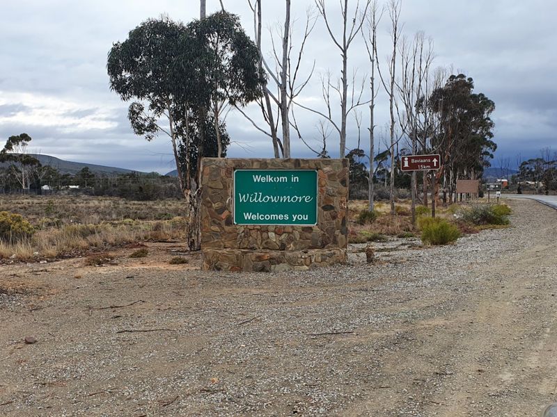 A street-level view of the Willowmore town entrance sign, featuring stone-clad masonry set against a rural landscape and adjacent asphalt road.
